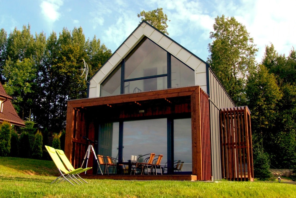 Nowoczesny dom z dużym przeszkleniem szczytowym i drewnianą pergolą, zieleń w tle, dwa zielone leżaki na trawniku.