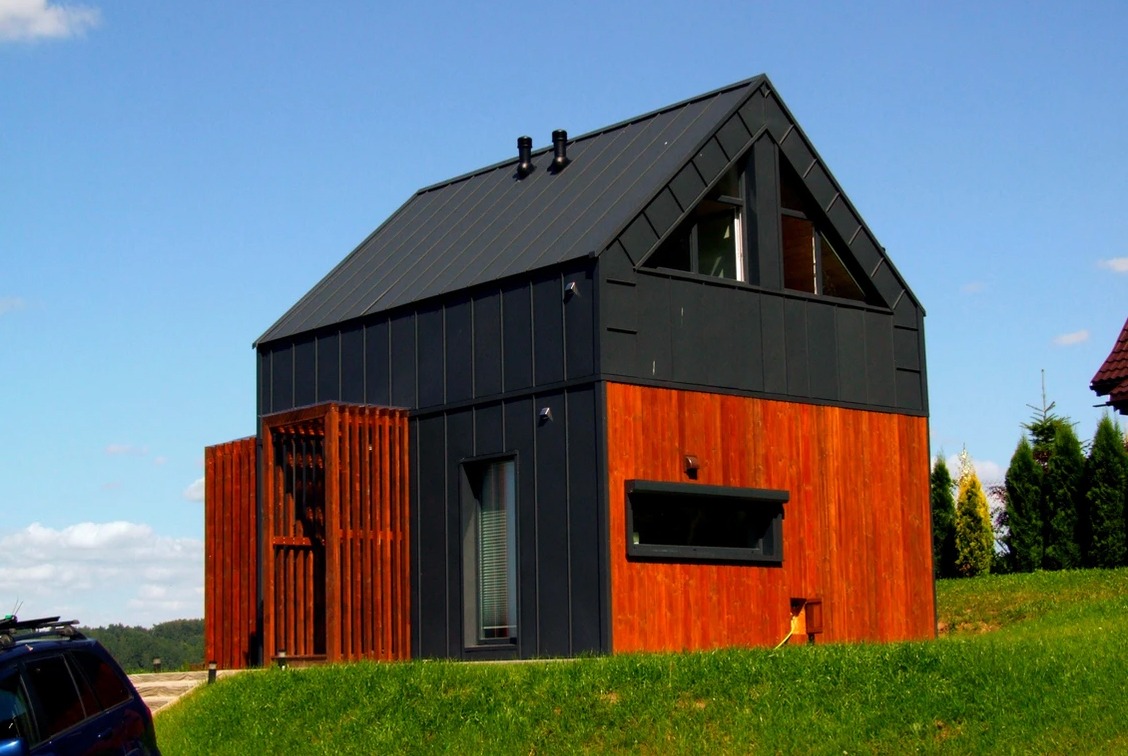 Nowoczesny dom z ciemnoszarą blachą na dachu i górnej części elewacji, z drewnianą dolną częścią i drewnianą ażurową konstrukcją wejściową, na tle zielonego trawnika i błękitnego nieba.