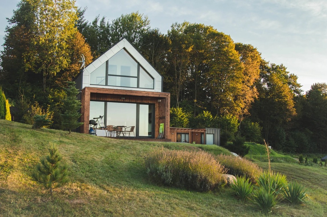 Nowoczesny dwukondygnacyjny dom z dużymi przeszkleniami, drewnianą elewacją i metalowym dachem, położony na wzgórzu z zielonym trawnikiem i drzewami w tle.