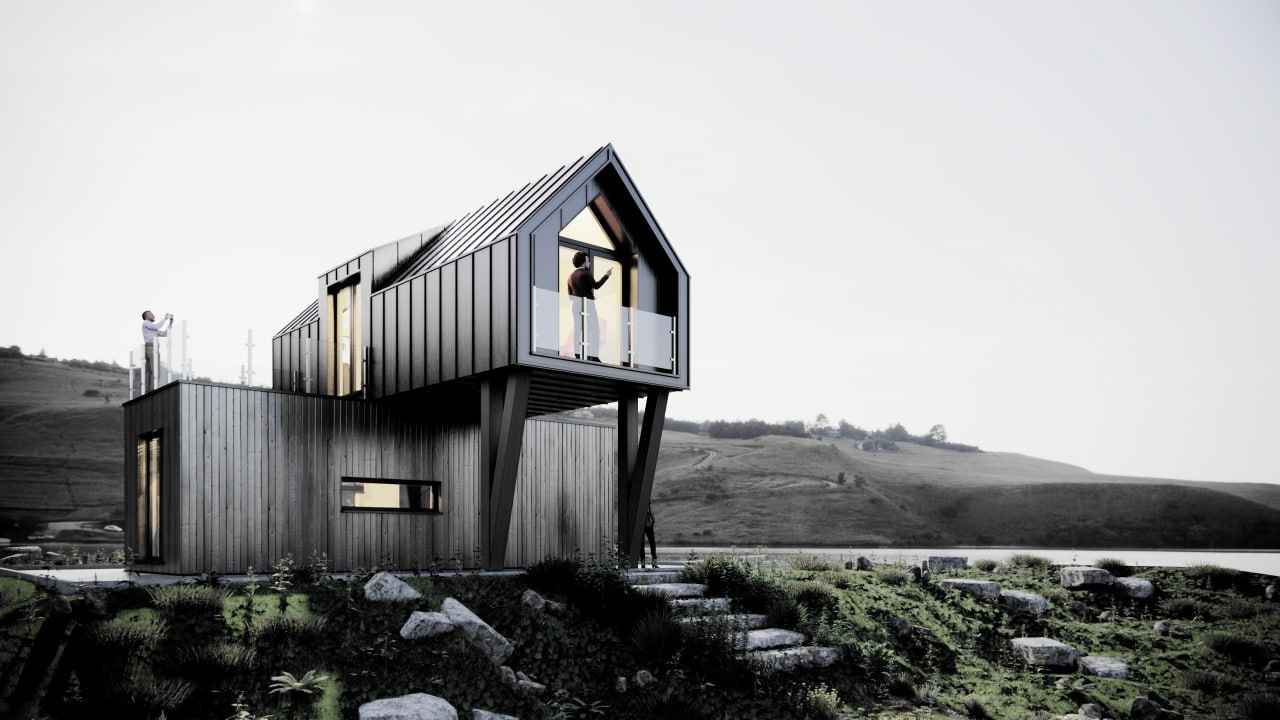 Czarno-drewniany dom modułowy z balkonem na wzgórzu, osoba na balkonie i druga na dachu robią zdjęcia otoczenia.