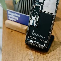 iHospital - Otwarty iPhone z widoczną baterią i chipem A15 Bionic, leżący na drewnianym biurku obok wizytówki i doniczki z rośliną.