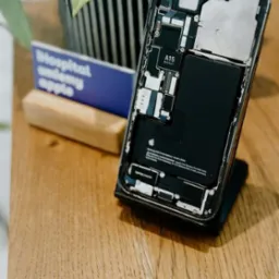 Otwarty iPhone z widoczną baterią i chipem A15 Bionic, leżący na drewnianym biurku obok wizytówki i doniczki z rośliną.