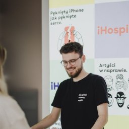 iHospital - Uśmiechnięty mężczyzna w okularach i czarnej koszulce z logo firmy iHospital obsługuje klienta przy biurku na tle plakatu z hasłem 'Pęknięty iPhone jak pęknięte serce.'