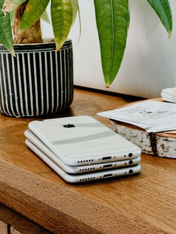 Stos trzech białych smartfonów iPhone leżących na drewnianym blacie, obok doniczki z rośliną i pliku kartek, widoczne porty ładowania i głośniki.