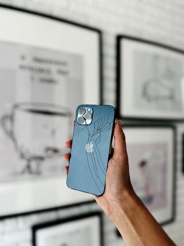 Niebieski smartfon z wyraźnie pękniętą tylną obudową, trzymany w dłoni na tle ściany z obrazami w czarnych ramkach.