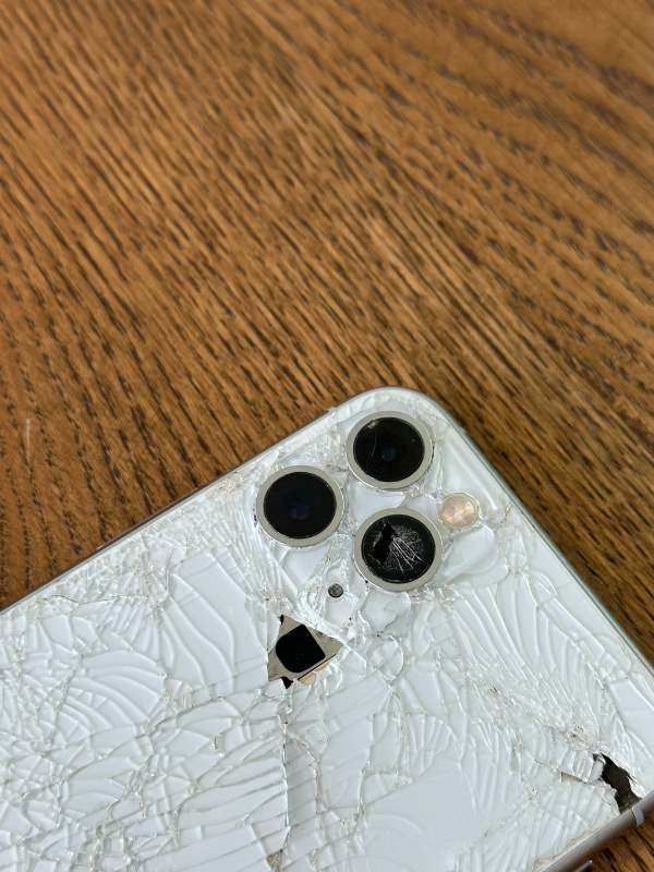 Smartfon z pękniętą, białą obudową i uszkodzonym obiektywem aparatu na drewnianym stole.