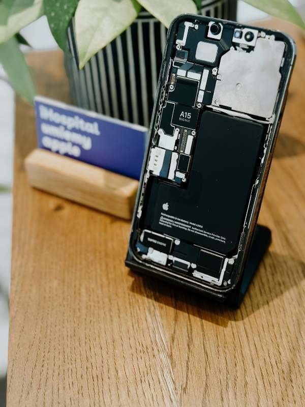 Otwarty iPhone z widoczną baterią i chipem A15 Bionic, leżący na drewnianym biurku obok wizytówki i doniczki z rośliną.