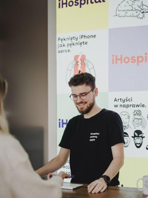 Uśmiechnięty mężczyzna w okularach i czarnej koszulce z logo firmy iHospital obsługuje klienta przy biurku na tle plakatu z hasłem 'Pęknięty iPhone jak pęknięte serce.'