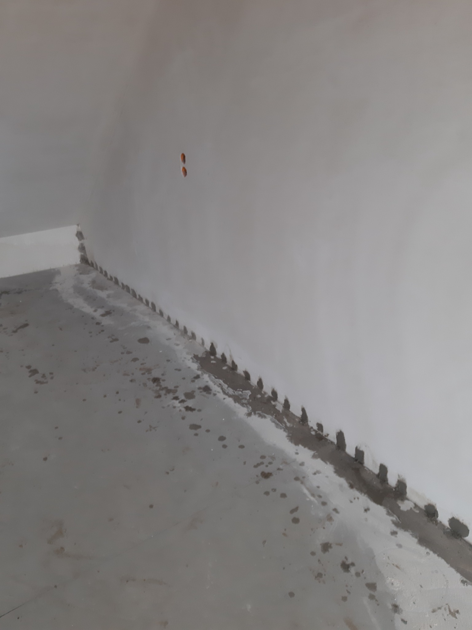 Szara podłoga z widocznymi plamami wilgoci i śladami po wierceniu otworów przy ścianie, przygotowana do procesu osuszania.