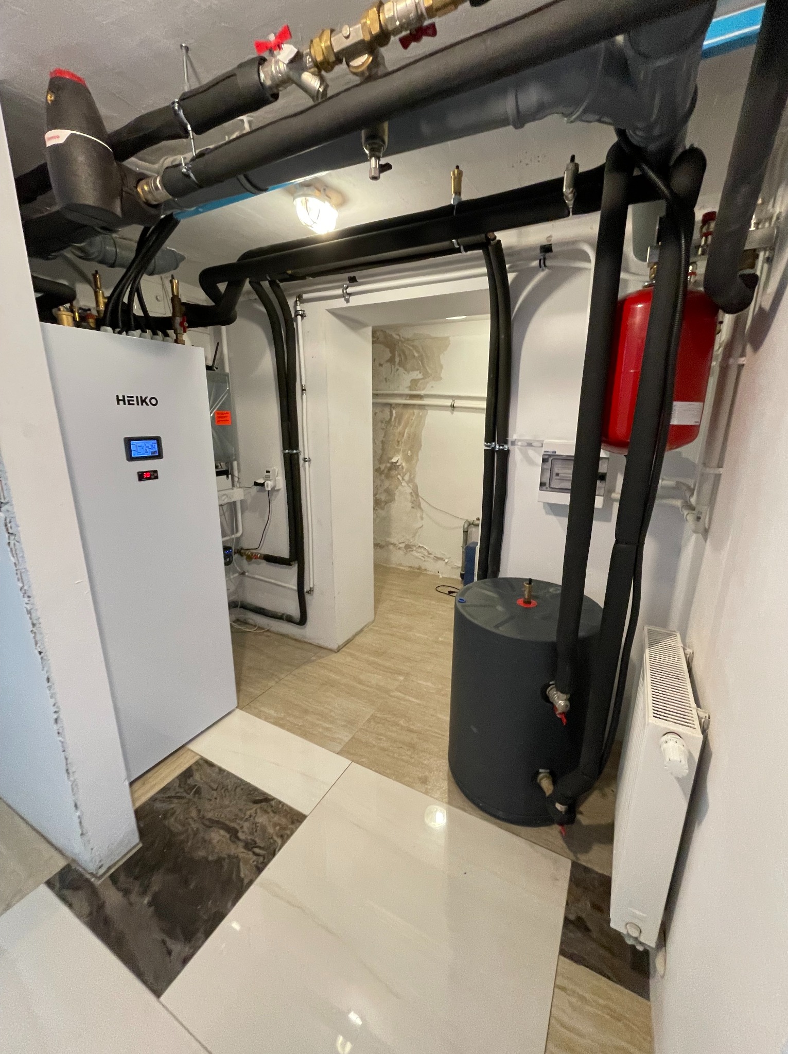 Kotłownia z pompą ciepła powietrze-woda HEIKO, zbiornikiem akumulacyjnym i grzejnikiem. Rury instalacji grzewczej z izolacją termiczną. W tle widoczne wejście do pomieszczenia.