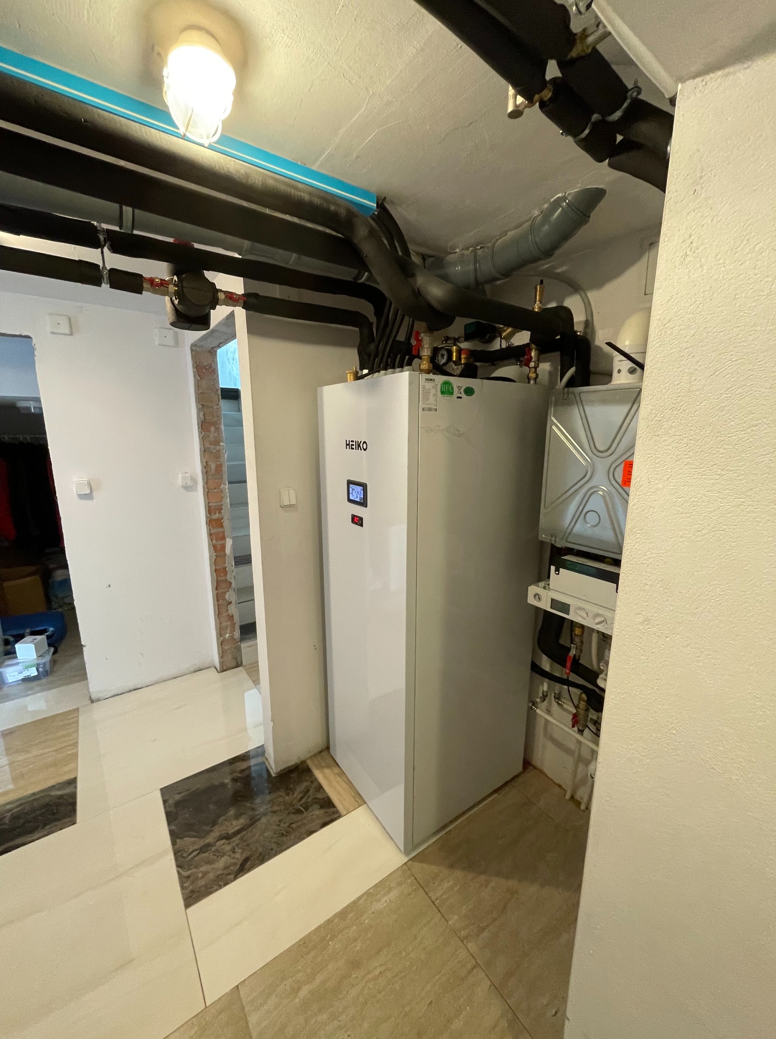 Wnętrze kotłowni z białą pompą ciepła HEIKO, widoczne rury instalacyjne pod sufitem i elementy montażowe na ścianie. Ograniczona przestrzeń, jasne oświetlenie.