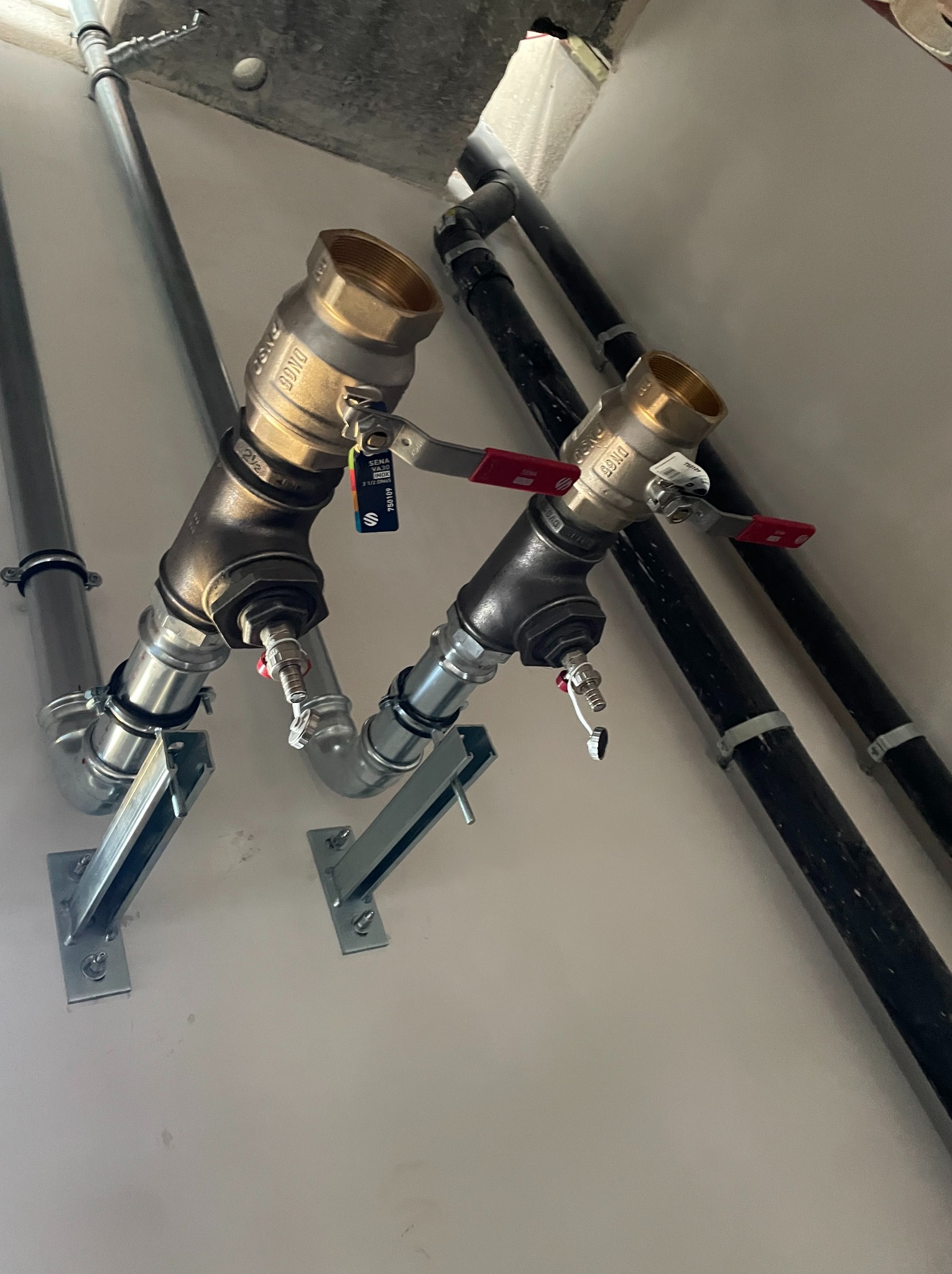 Dwie pionowe rury z zaworami kulowymi, wsparte na metalowych wspornikach, instalacja w hali produkcyjnej. Rury szare i czarne, zawory mosiężne z czerwonymi uchwytami.