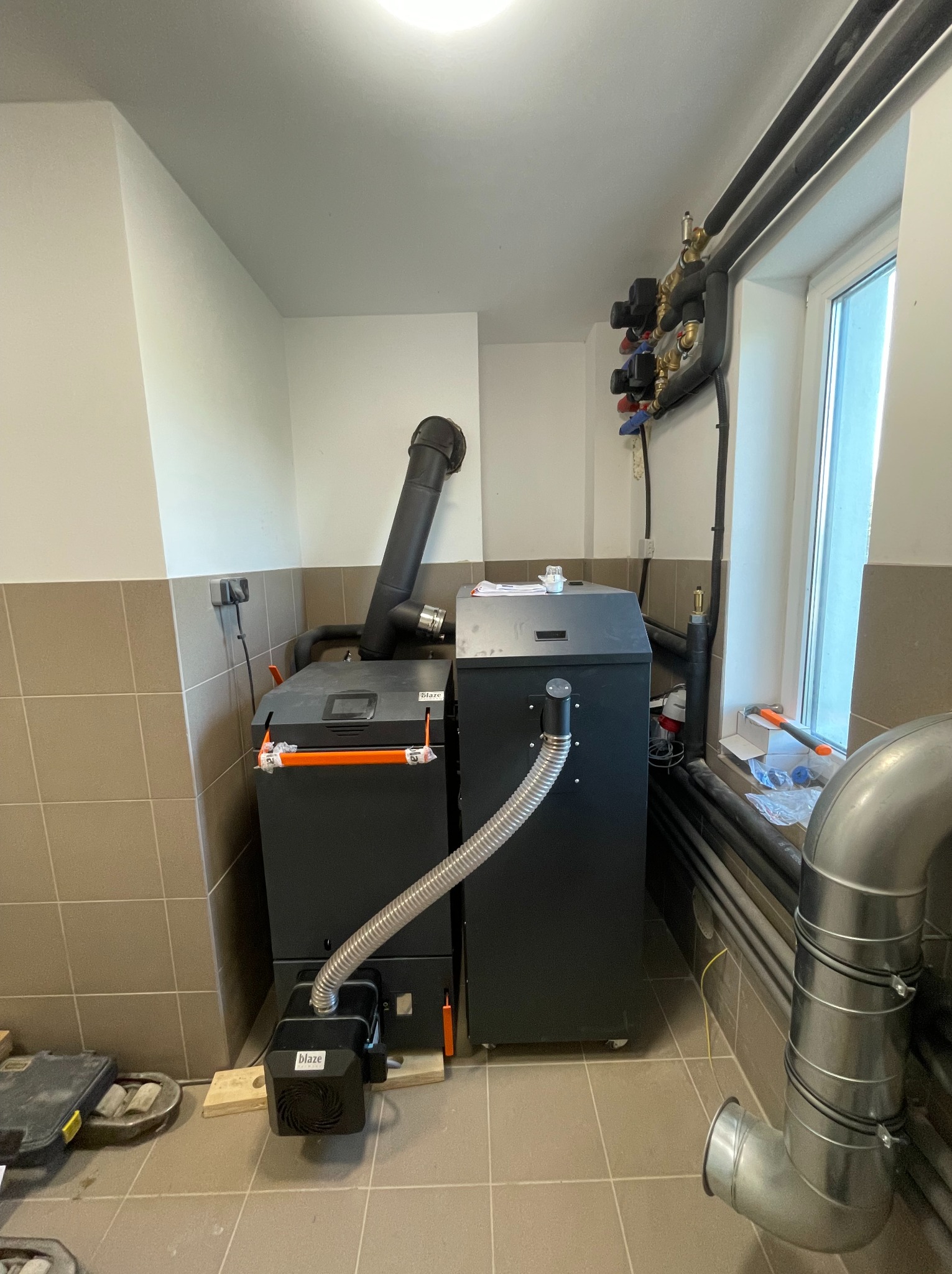 Kocioł kombi zgazowujący drewno i pellet w kotłowni domu jednorodzinnego. Widoczne rury, okno i płytki na ścianach. Kocioł marki Blaze.