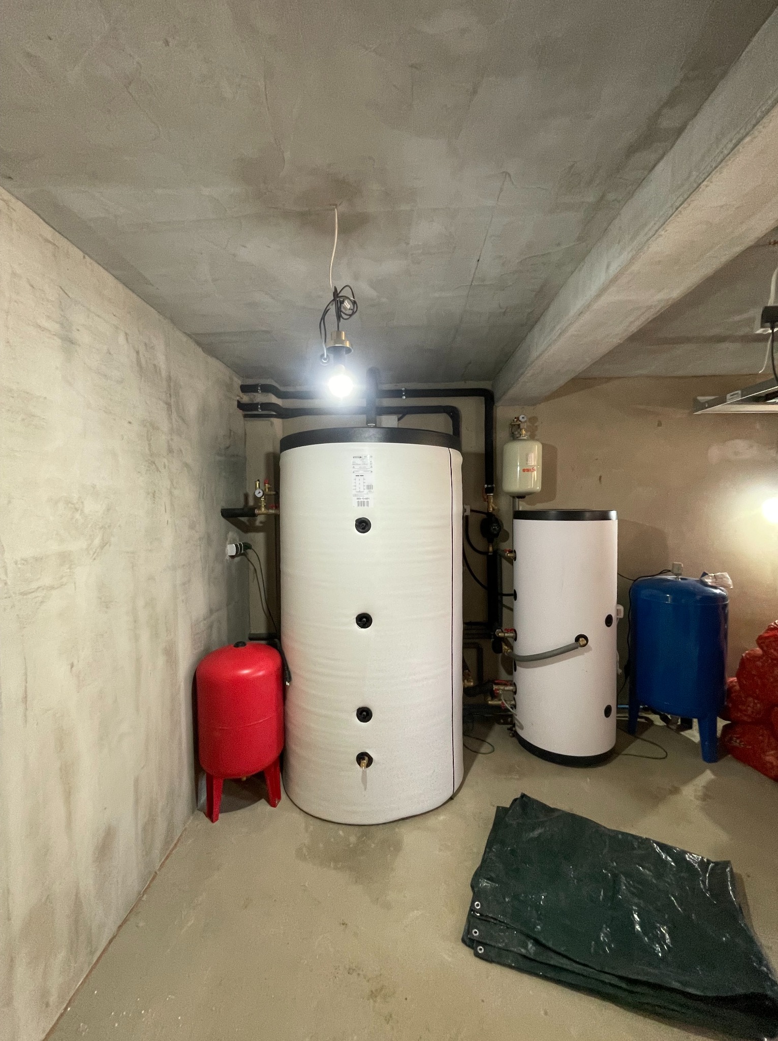 Kotłownia z kotłem kombi w Kwidzynie. Widoczne zbiorniki, rury i osprzęt. Surowy beton na ścianach i suficie. Instalacja połączona, gotowa do użytku.