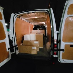 GWRD ANNA KACZMAREK - Wnętrze białego busa z otwartymi tylnymi drzwiami, załadowanego kartonami o różnych rozmiarach, widok z tyłu w słabym oświetleniu.