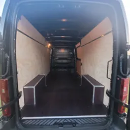 Wnętrze czarnego busa z wyłożonymi sklejką ścianami i dwiema skrzyniami po bokach, widok z tyłu przy otwartych drzwiach.