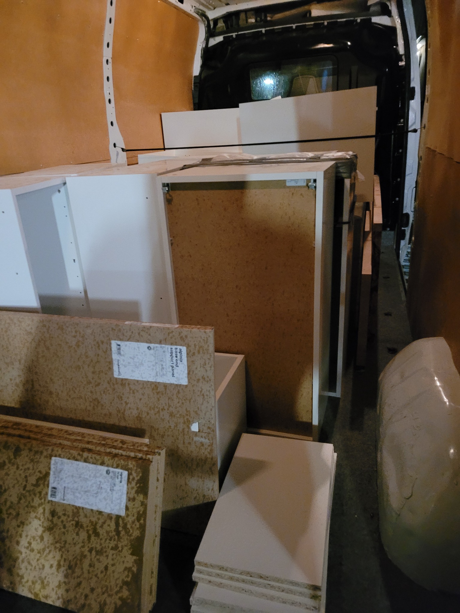 Wnętrze furgonetki załadowane elementami meblowymi, w tym płytami wiórowymi z etykietami i białymi szafkami, zabezpieczonymi do transportu.