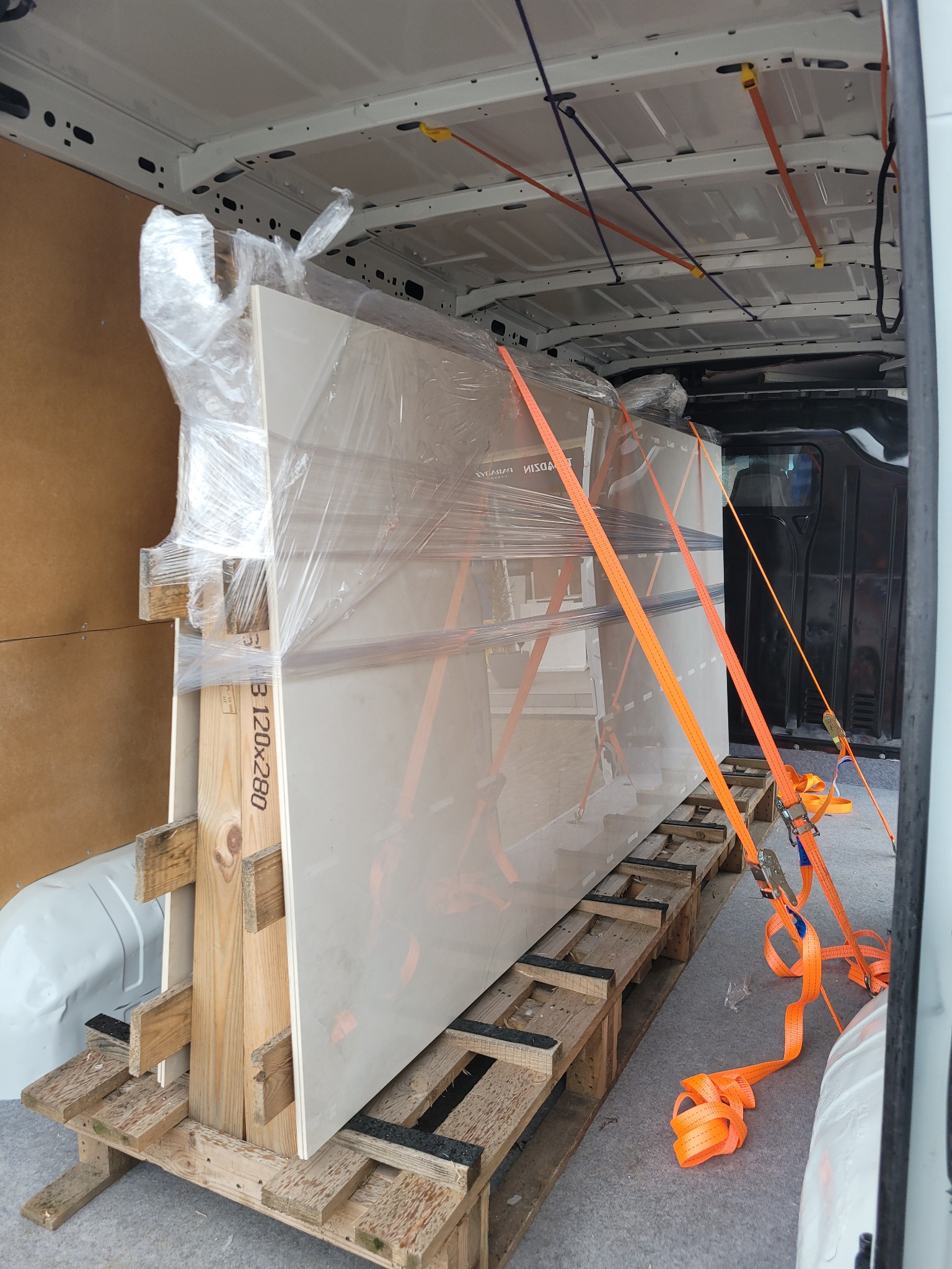 Płyta ceramiczna zabezpieczona folią i pasami transportowymi na palecie wewnątrz furgonetki, widoczny fragment etykiety B 120x280.