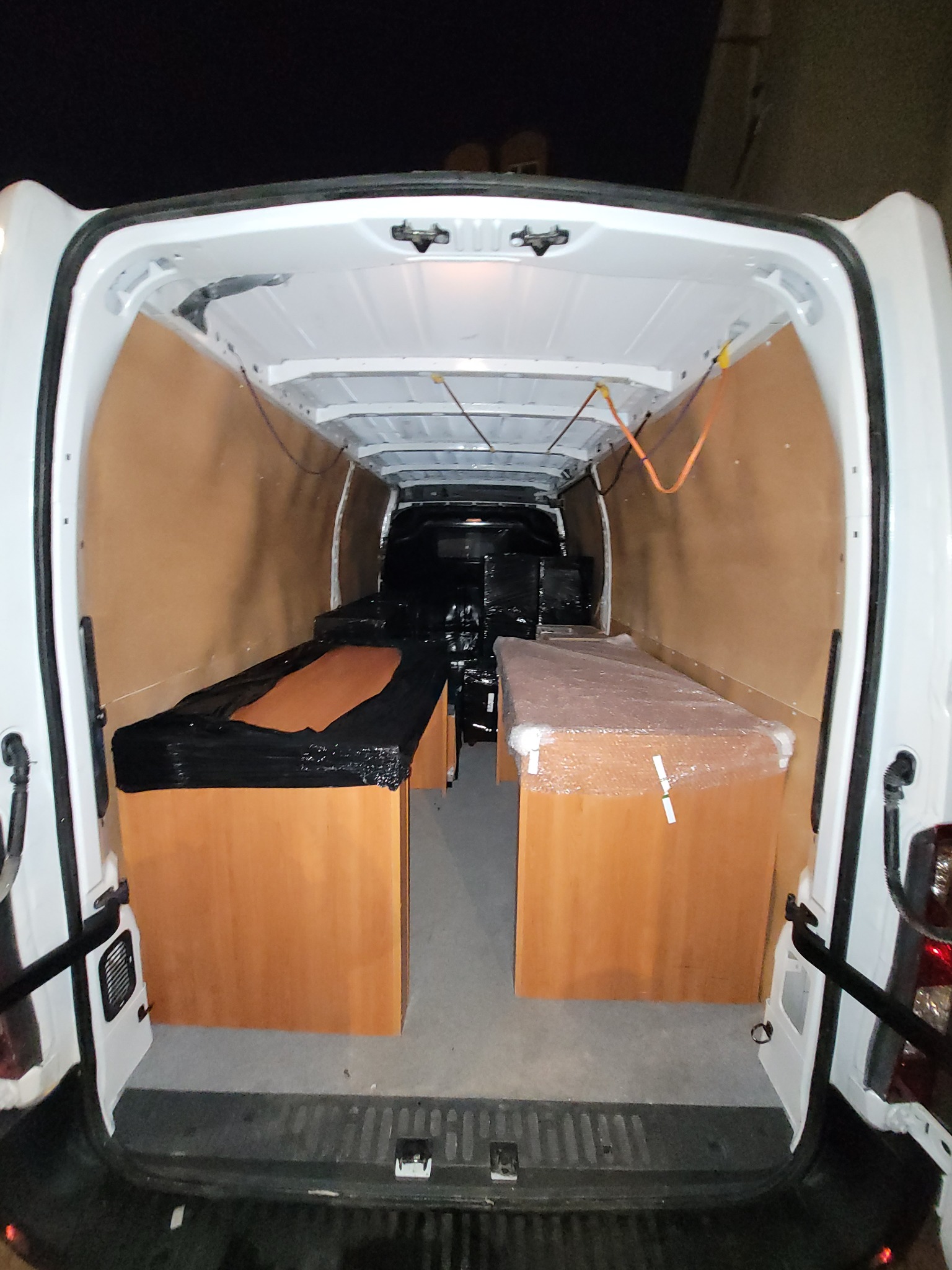 Wnętrze białego furgonetki wyłożone sklejką, załadowane biurkami zabezpieczonymi folią i czarną folią stretch, gotowe do transportu.