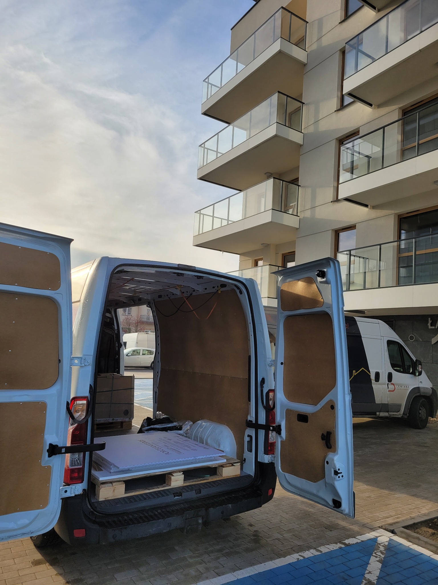 Otwarty furgon z paletą i kartonami na tle nowoczesnego apartamentowca z balkonami, drugi furgon widoczny w tle, poranne oświetlenie.