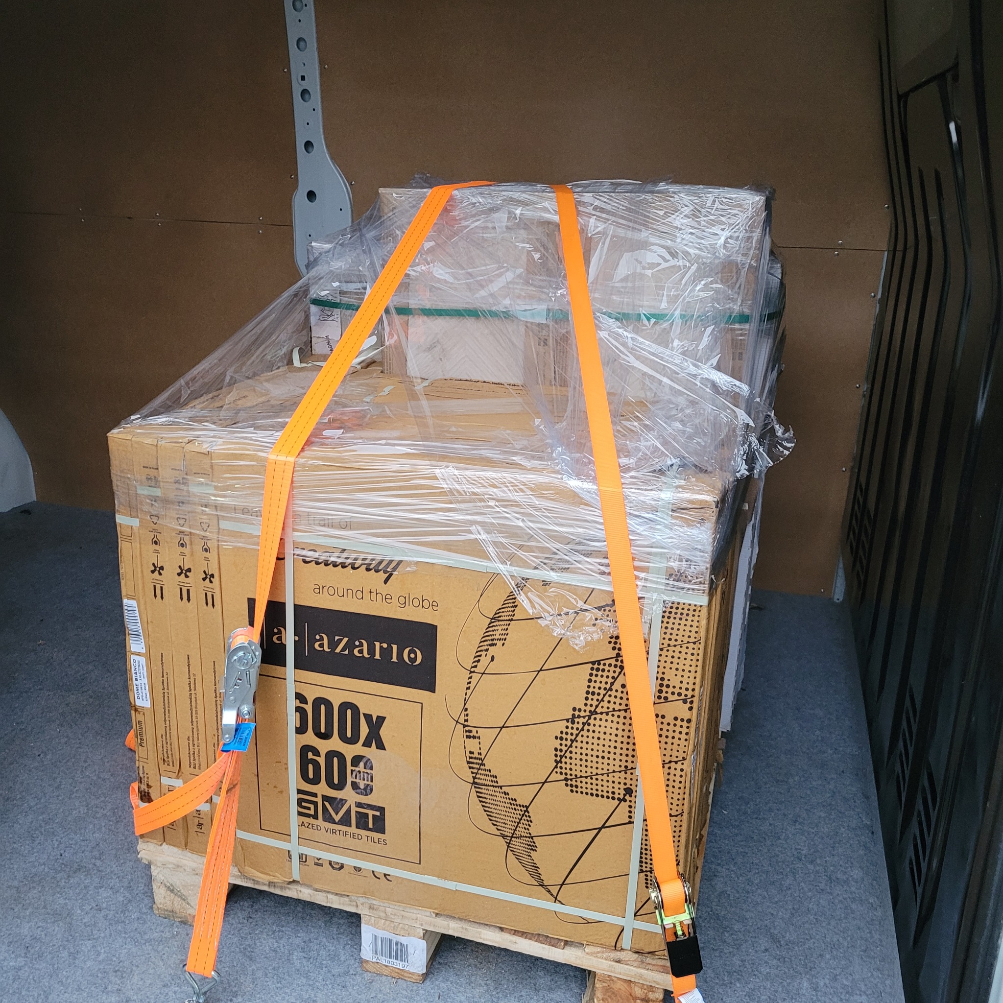 Paleta z kartonami płytek, zabezpieczona folią i pomarańczowymi pasami transportowymi, wewnątrz przestrzeni ładunkowej pojazdu.