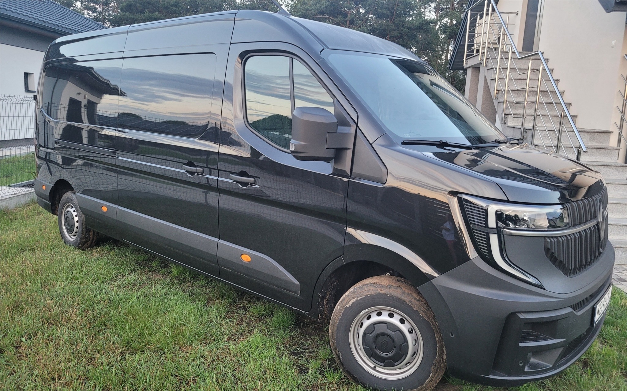 Czarny, nowoczesny bus dostawczy zaparkowany na trawie przed domem. Widoczne detale karoserii i oświetlenia.