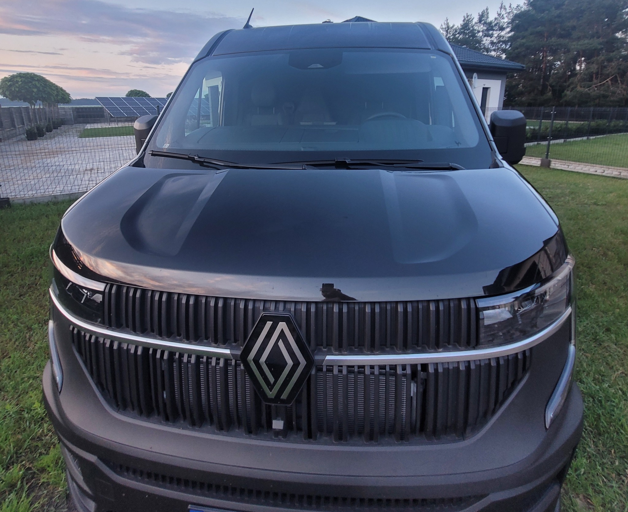 Czarny bus Renault z przodu, z widocznym grillem i logo, na tle domu i zieleni. Ujęcie z bliska podkreśla detale pojazdu.