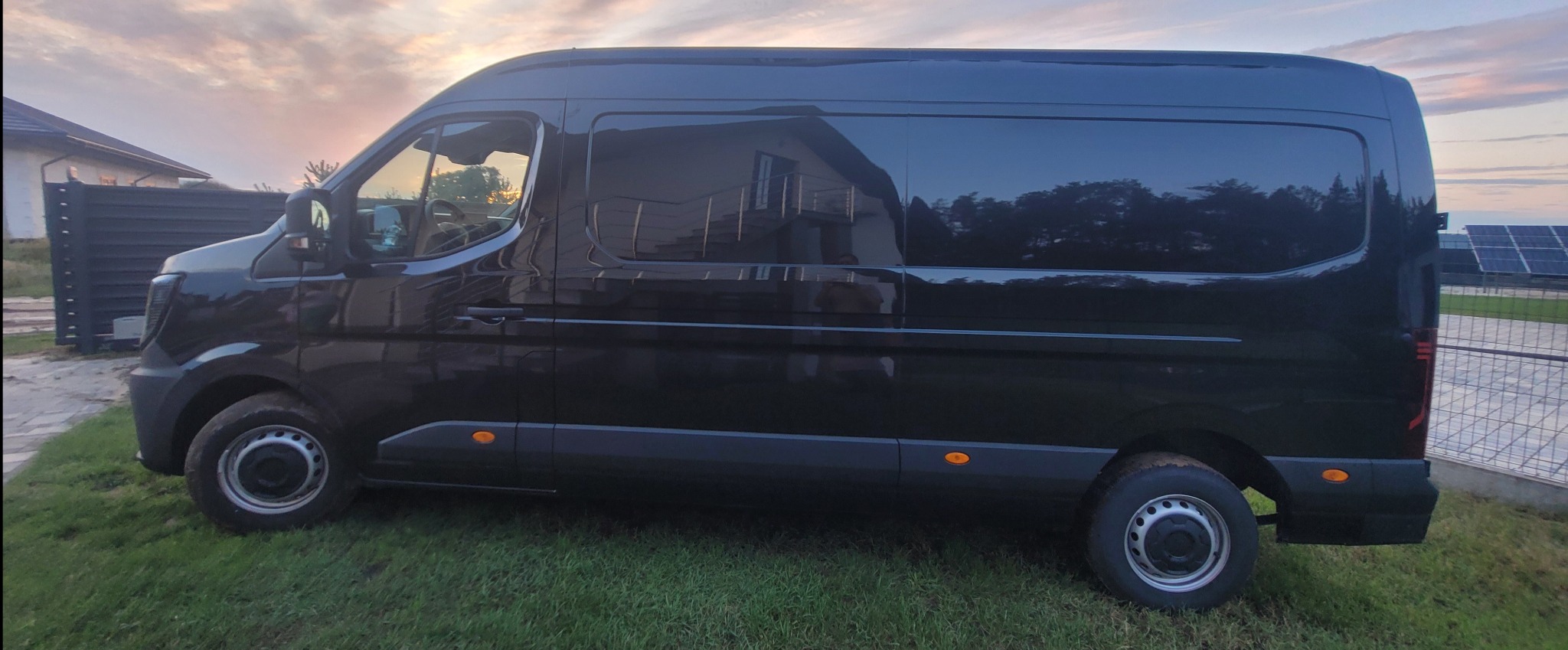 Czarny, lśniący bus na trawie z odbiciem domu w szybach, w tle zachodzące słońce i panele słoneczne. Widok z boku, pokazujący całą sylwetkę pojazdu.
