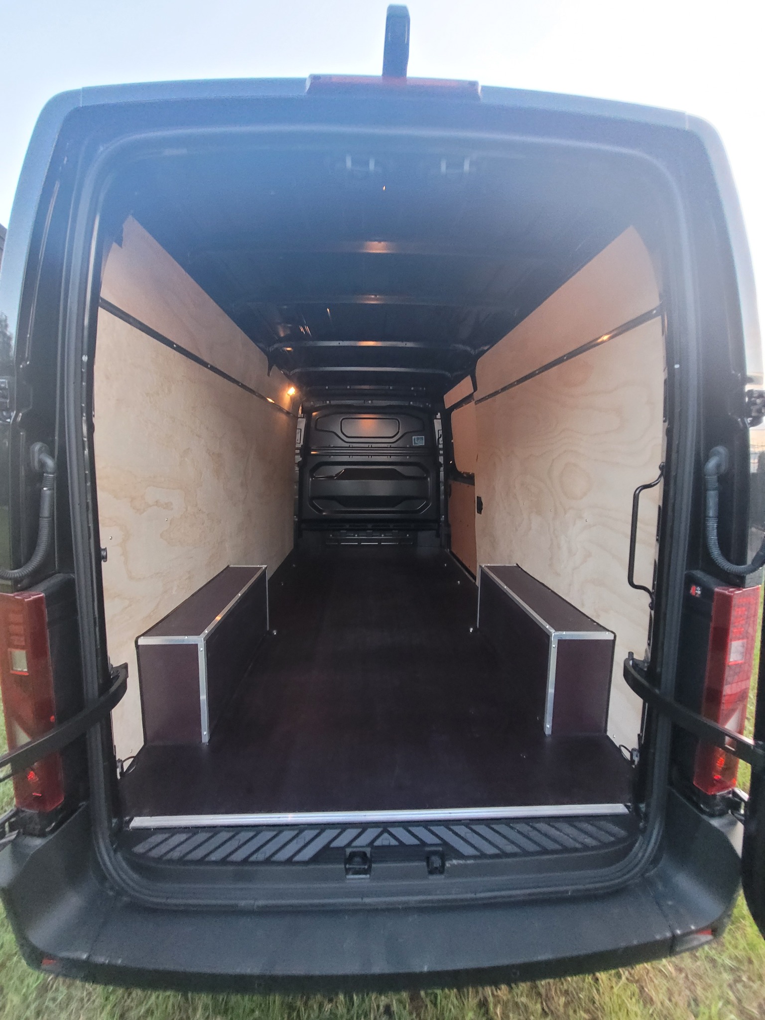 Wnętrze czarnego busa z wyłożonymi sklejką ścianami i dwiema skrzyniami po bokach, widok z tyłu przy otwartych drzwiach.