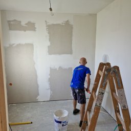 AS-BUD DAMIAN KANIA - Mężczyzna w niebieskiej koszulce wyrównuje ścianę szpachelką w pomieszczeniu z surowym oświetleniem, widoczna drabina, wiadro z masą szpachlową i narzędzia.
