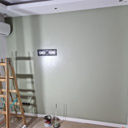 AS-BUD DAMIAN KANIA - Ściana w odcieniach zieleni z delikatnym wzorem jodełki, uchwyt do telewizora, drabina malarska i akcesoria malarskie na podłodze.