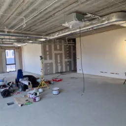 Wnętrze biura w trakcie remontu: surowy betonowy sufit, instalacje, ściany przygotowane do malowania, narzędzia i materiały malarskie. Oświetlenie budowlane.