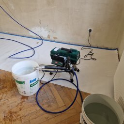 AS-BUD DAMIAN KANIA - Przygotowanie do natryskowego nakładania gładzi: wiadro z masą Knauf, agregat malarski, zabezpieczona podłoga i ściany taśmą malarską.