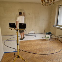 AS-BUD DAMIAN KANIA - Pracownik szpachluje ścianę w pokoju z parkietem, używając agregatu. Podłoga zabezpieczona folią, okno zaklejone, widoczne lampy oświetleniowe i wiadro z masą.