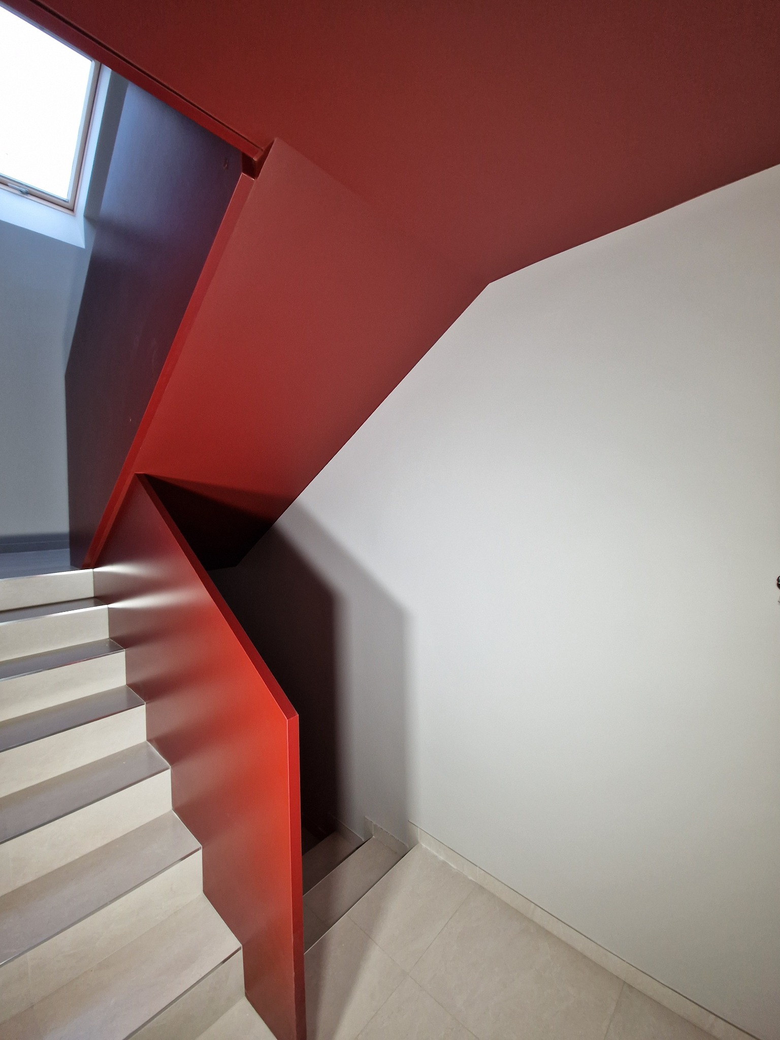Nowoczesne schody z czerwonymi elementami pomalowanymi na intensywny odcień czerwieni, kontrastujące z białą ścianą i jasnymi stopniami, oświetlone światłem wpadającym przez okno dachowe.