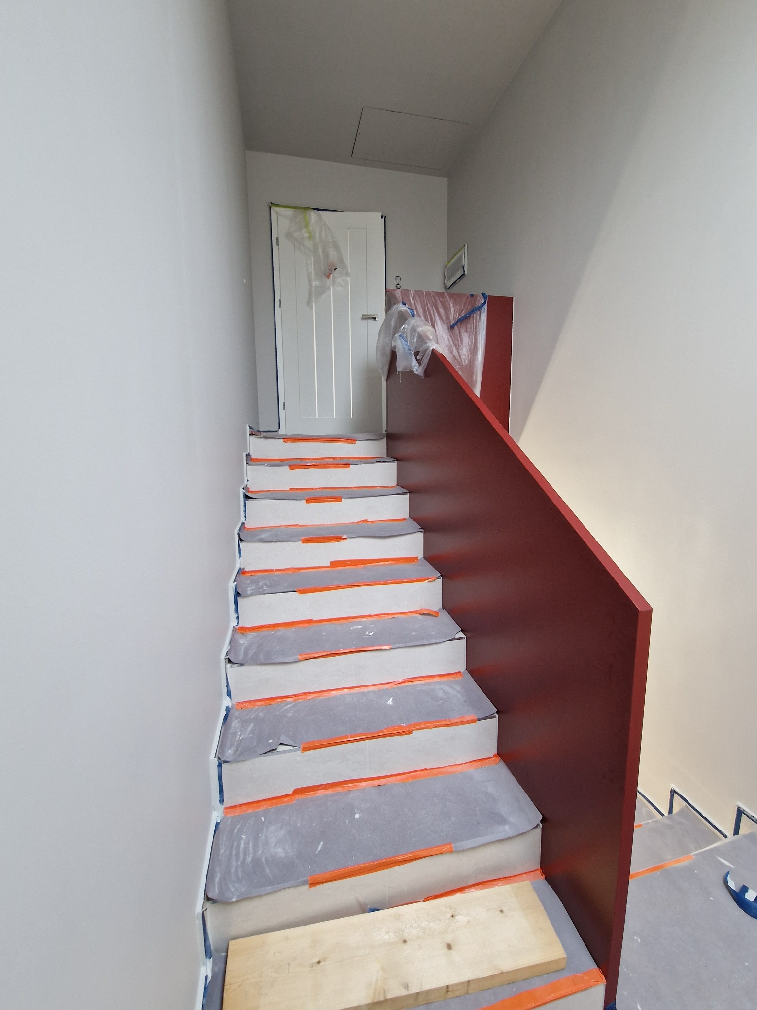 Schody w trakcie remontu: zabezpieczone folią malarską stopnie, świeżo pomalowana ściana na szaro, bordowa balustrada. Widoczny element drewniany na dolnym stopniu.