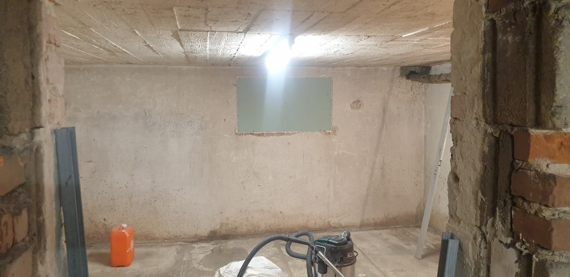 Surowy pokój w trakcie prac przygotowawczych: fragment ściany z zamontowaną płytą gipsowo-kartonową, widoczne ślady po obróbce murarskiej na suficie, odkurzacz przemysłowy na podłodze.