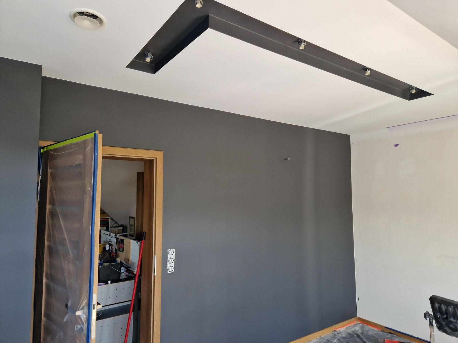 Pokój w trakcie odświeżania: pomalowana na szaro ściana z widoczną instalacją oświetleniową na suficie, zabezpieczone folią drzwi i fragmenty ścian przygotowywane do dalszych prac.