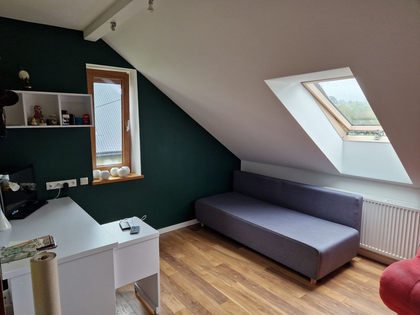 Pokój na poddaszu z ciemnozieloną ścianą, białym biurkiem i szarą sofą pod oknem dachowym; widoczne drewniane panele podłogowe.