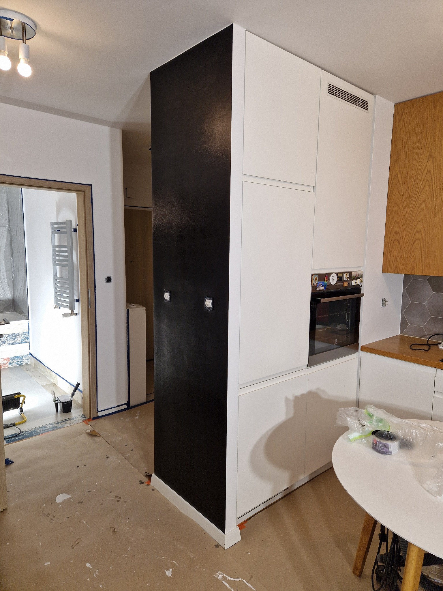 Wnętrze mieszkania w trakcie remontu: czarna ściana po malowaniu, zabezpieczona podłoga, białe meble kuchenne z piekarnikiem, widoczne akcesoria malarskie.