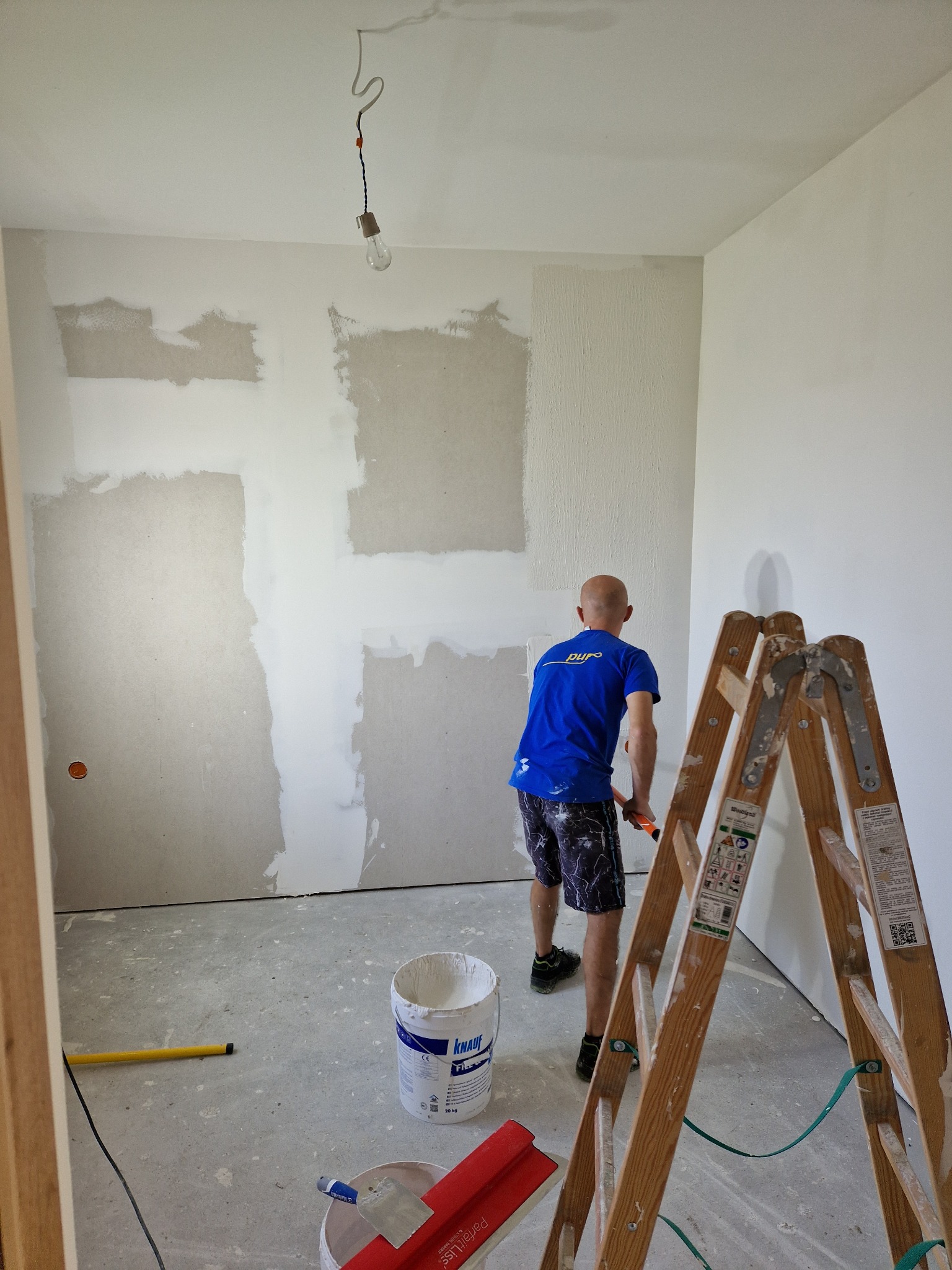Mężczyzna w niebieskiej koszulce wyrównuje ścianę szpachelką w pomieszczeniu z surowym oświetleniem, widoczna drabina, wiadro z masą szpachlową i narzędzia.