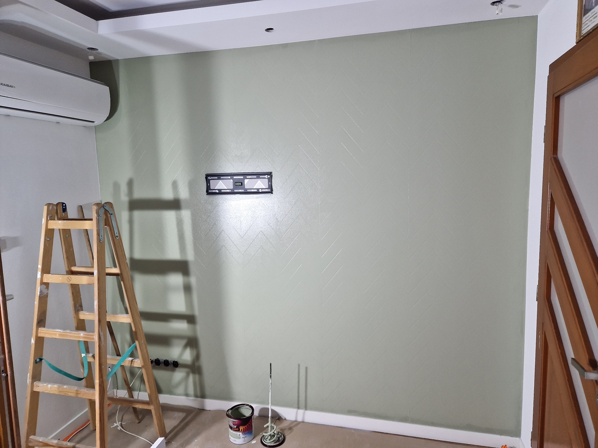 Ściana w odcieniach zieleni z delikatnym wzorem jodełki, uchwyt do telewizora, drabina malarska i akcesoria malarskie na podłodze.