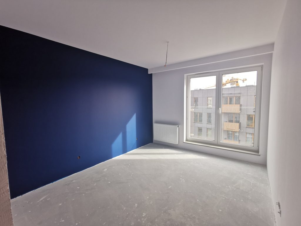 Wnętrze pokoju po częściowym remoncie: jedna ściana pomalowana na ciemny granat, reszta ścian w odcieniach szarości, podłoga surowa, widoczne okno z widokiem na budynek w budowie.