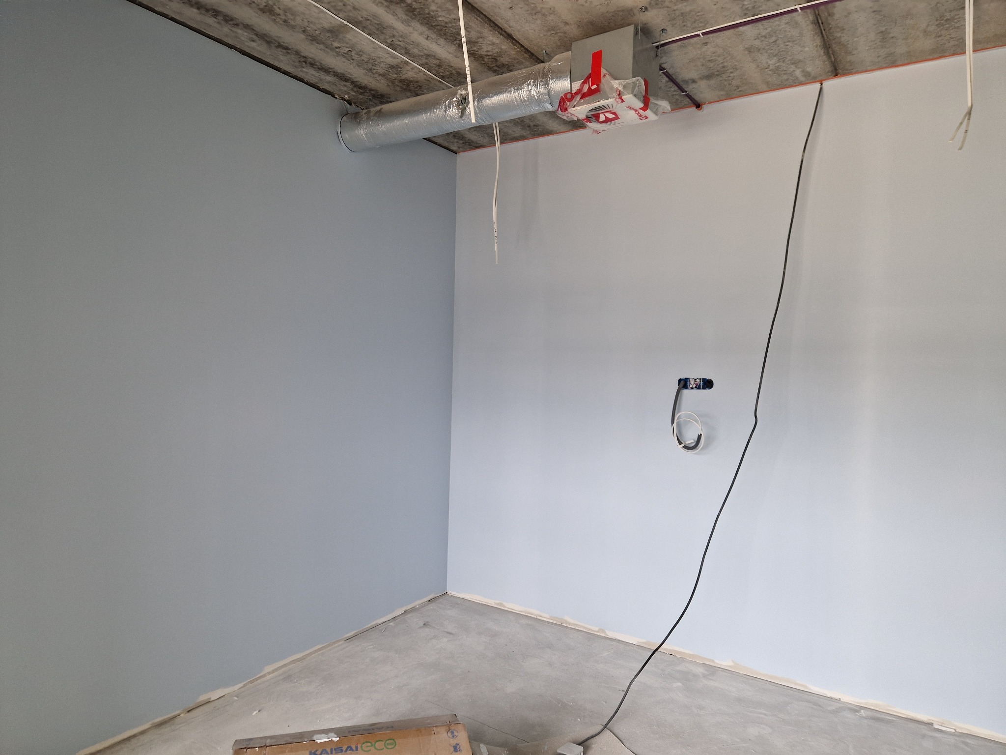 Wnętrze po malowaniu ścian na jasny odcień szarości, widoczne instalacje pod sufitem i gniazdko elektryczne. Na podłodze karton.