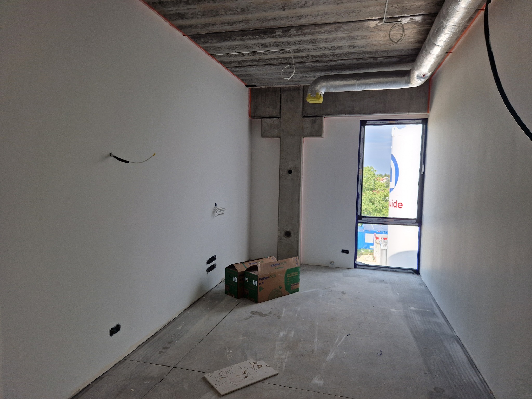 Surowe wnętrze po szpachlowaniu: gładkie, białe ściany, betonowy sufit z instalacją, okno z widokiem na zieleń. Na podłodze kartony i płyta. Widoczne puszki elektryczne.
