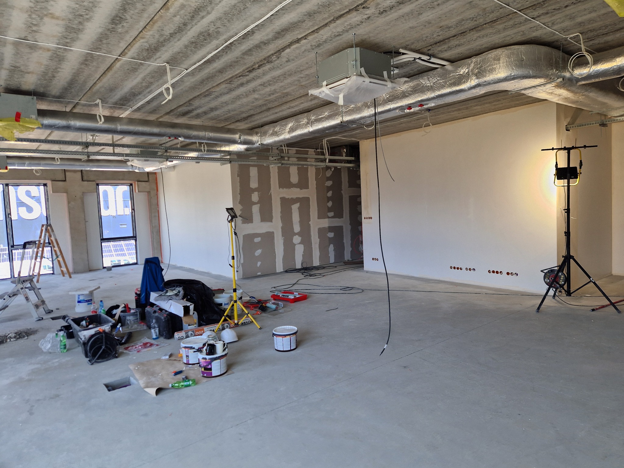 Wnętrze biura w trakcie remontu: surowy betonowy sufit, instalacje, ściany przygotowane do malowania, narzędzia i materiały malarskie. Oświetlenie budowlane.