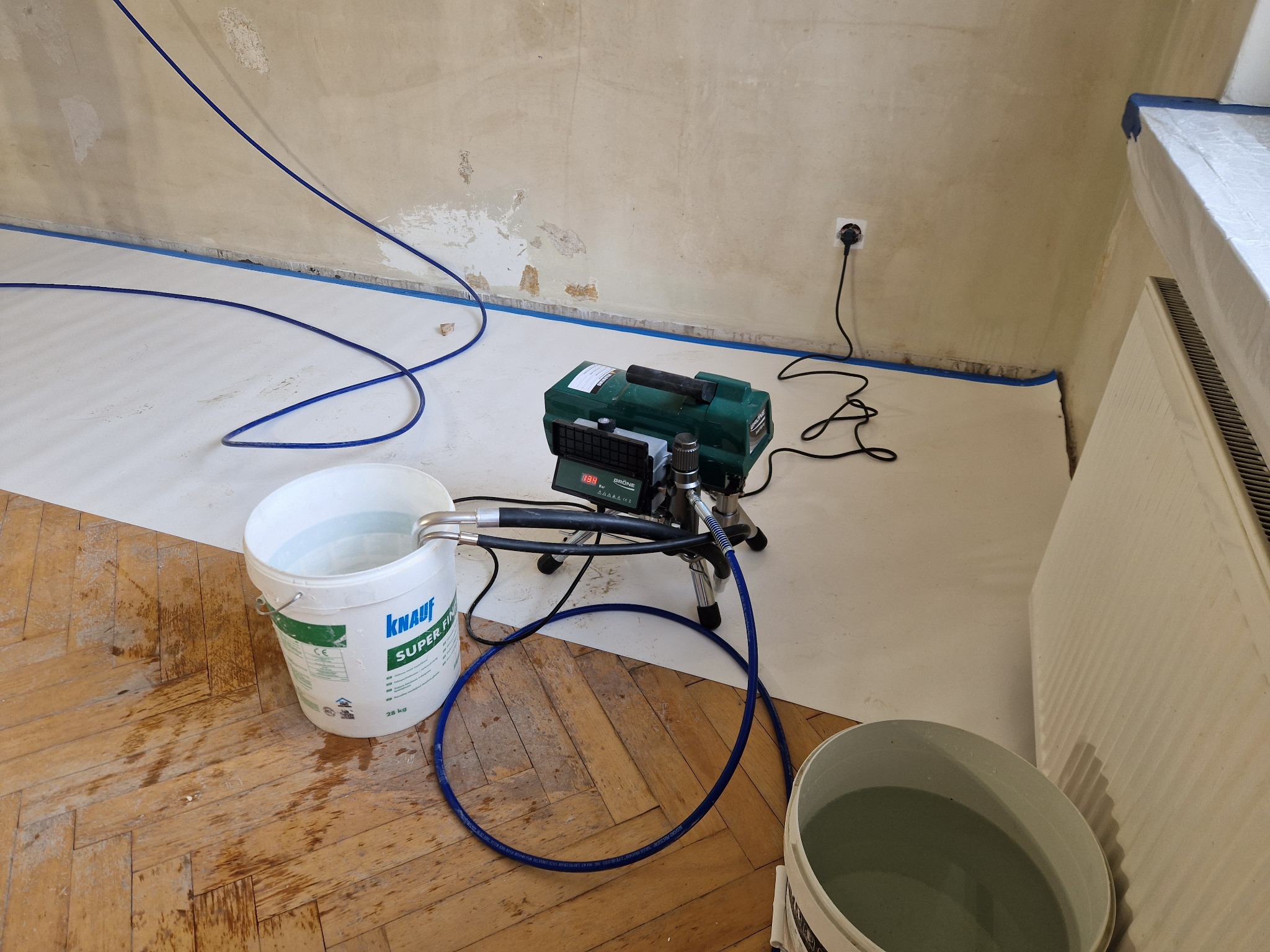 Przygotowanie do natryskowego nakładania gładzi: wiadro z masą Knauf, agregat malarski, zabezpieczona podłoga i ściany taśmą malarską.