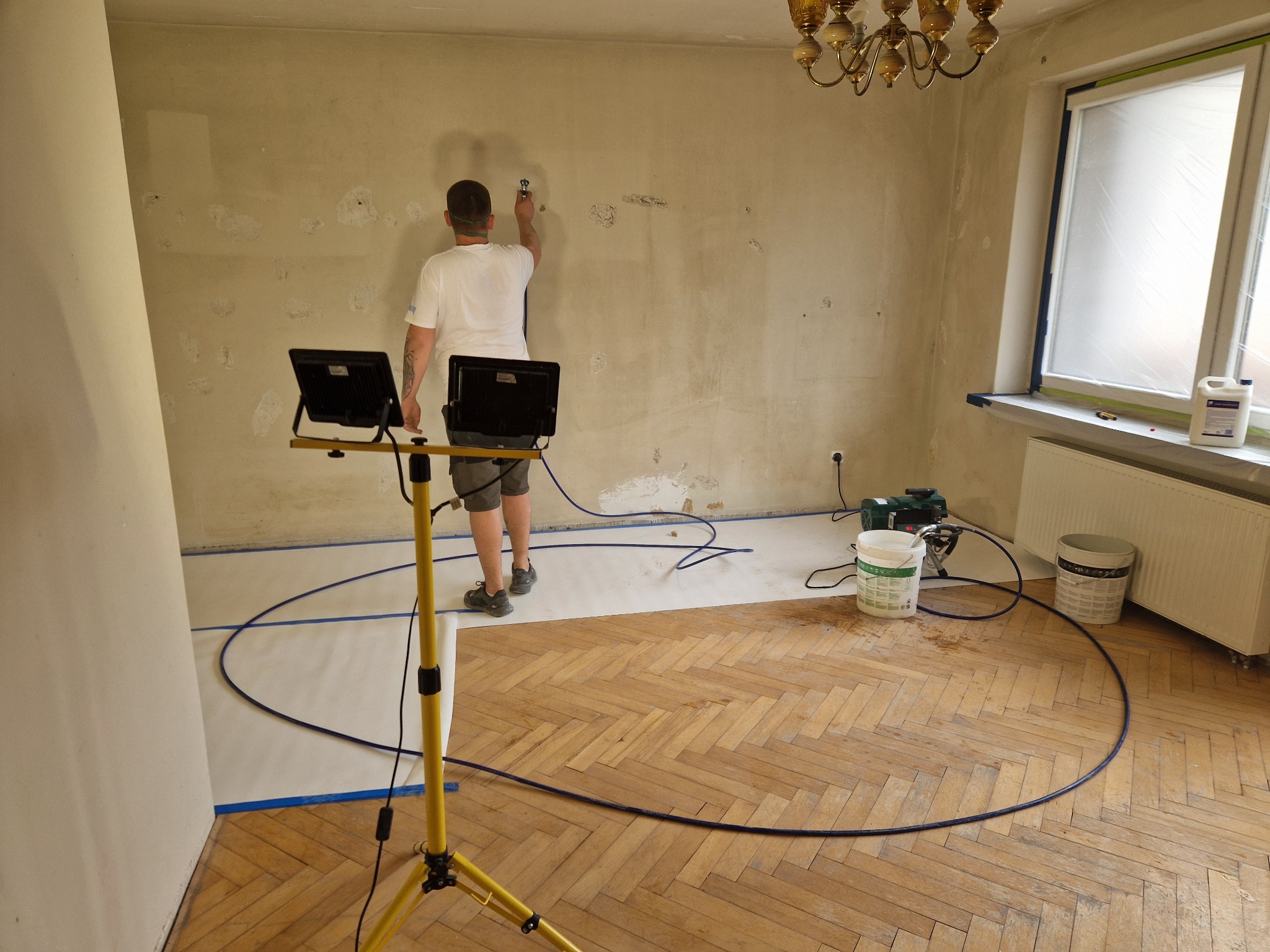 Pracownik szpachluje ścianę w pokoju z parkietem, używając agregatu. Podłoga zabezpieczona folią, okno zaklejone, widoczne lampy oświetleniowe i wiadro z masą.