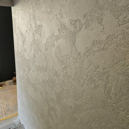 HYDBUD - Ściana pokryta dekoracyjnym tynkiem strukturalnym w odcieniach szarości, z widoczną fakturą i nieregularnym wzorem, w tle fragment czarnego otworu drzwiowego i zabezpieczona podłoga.