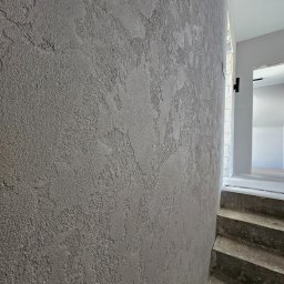 HYDBUD - Szara, teksturowana ściana z efektem dekoracyjnym, widoczna obok betonowych schodów w surowym stanie i fragmentu jasnego, wykończonego wnętrza w tle.